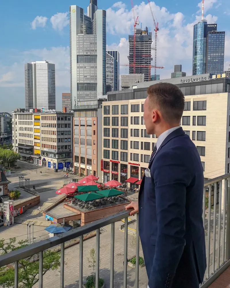 Arthur in a suit on a balcony above the Frankfurt skyline